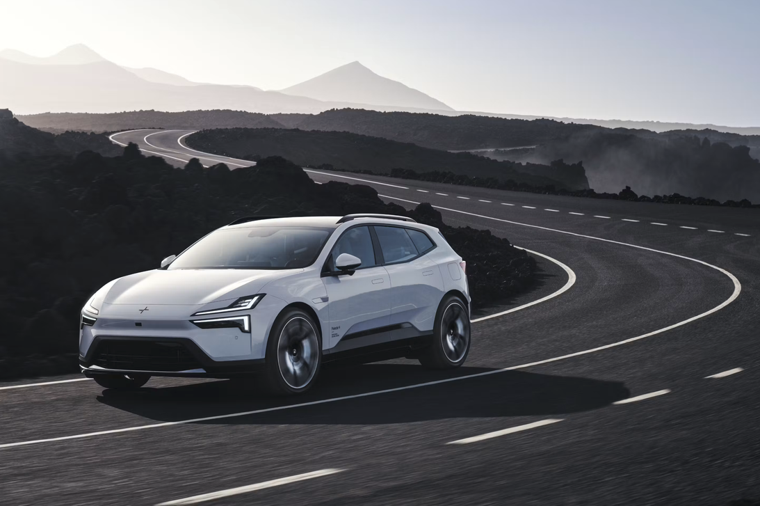 A white Polestar 4 SUV driving on a winding asphalt road through a volcanic, rocky landscape with hazy mountains visible in the distance under a pale sky.